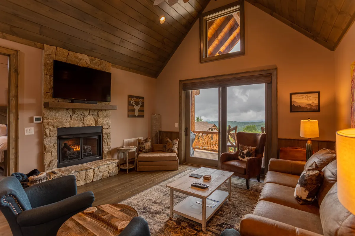 An inviting interior scene featuring a spacious living area with a cozy fireplace, large windows that flood the room with light, and comfortable seating arranged for conversation. The space is decorated with art and has a balcony visible, suggesting indoor-outdoor flow and a setting ideal for gatherings.