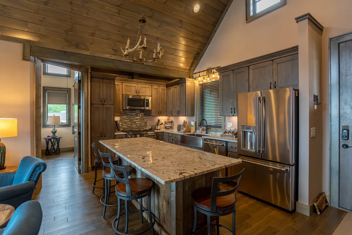This image focuses on a sleek kitchen space featuring a granite countertop, wooden cabinetry, and stylish bar stools arranged at the island. The warm lighting from a chandelier above adds to the cozy, welcoming atmosphere, inviting guests to gather around for conversations or meals.