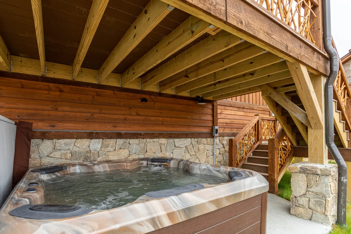 This exterior image features a bubbling hot tub nestled against a stone wall with sturdy wooden beams overhead, all set in a tranquil outdoor area. The hot tub is positioned to take in the surrounding natural beauty, promising relaxation and scenic enjoyment.