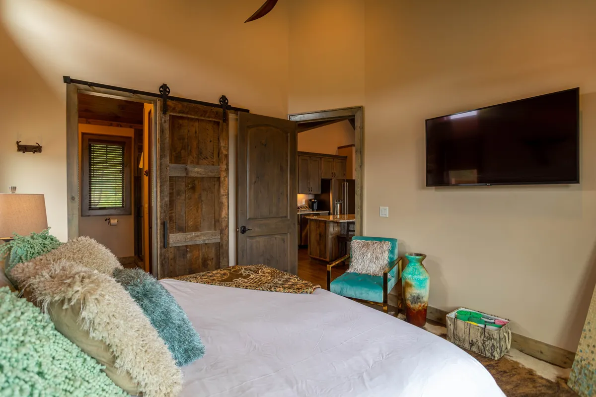A king suite bedroom featuring a bed with decorative pillows, sliding barn doors, a wall-mounted TV, a green accent chair, and a colorful vase, blending comfort and modern rustic style.