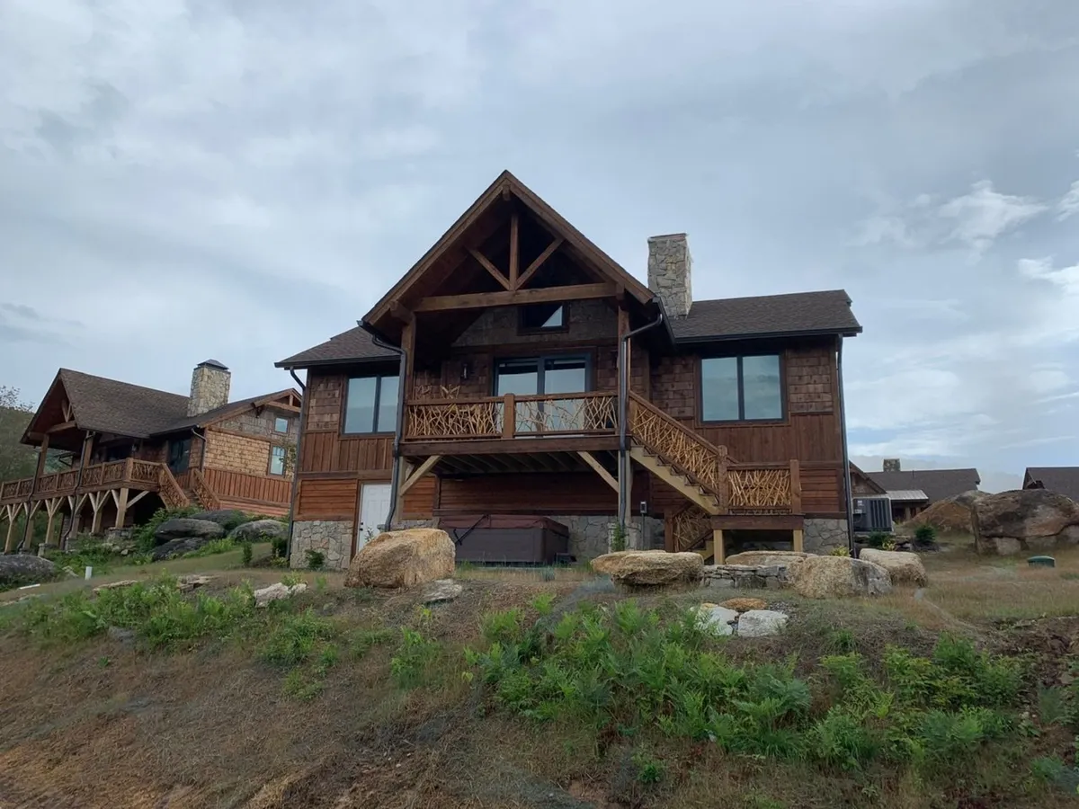 This view showcases a spacious wooden front porch with a sturdy stone chimney and a staircase leading to the entrance. The porch is designed for gatherings, offering a cozy spot to enjoy the outdoors and the landscape beyond.