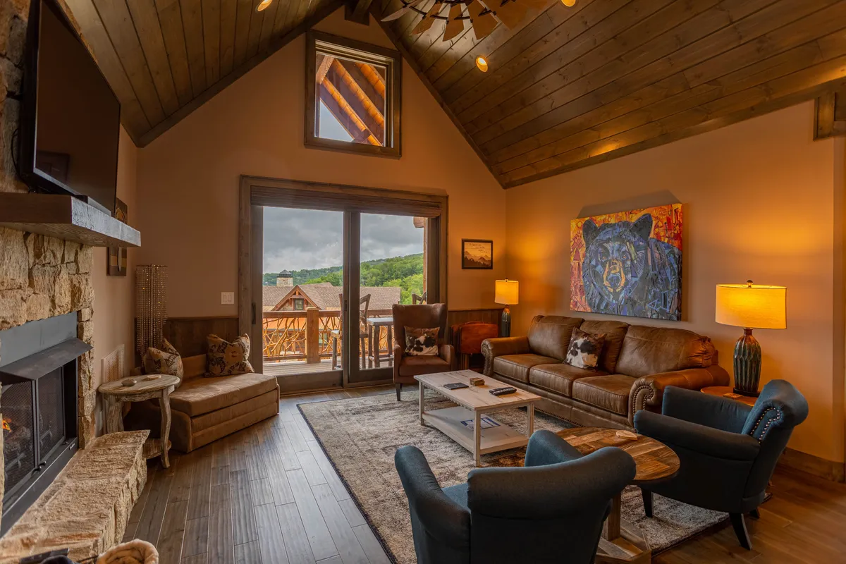 This image showcases a warm and inviting living room with a prominent gas fireplace, large windows that let in natural light, a leather sofa, and notable art featuring a bear. Wooden beams above add rustic charm, while the overall space feels open and connected to the outdoors.