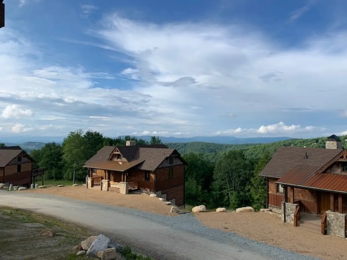 A panoramic exterior view of charming wooden cabins nestled against distant mountains beneath a cloudy sky. The scene is tranquil yet inviting, with hints of families and guests gathering, and paths leading further into nature.