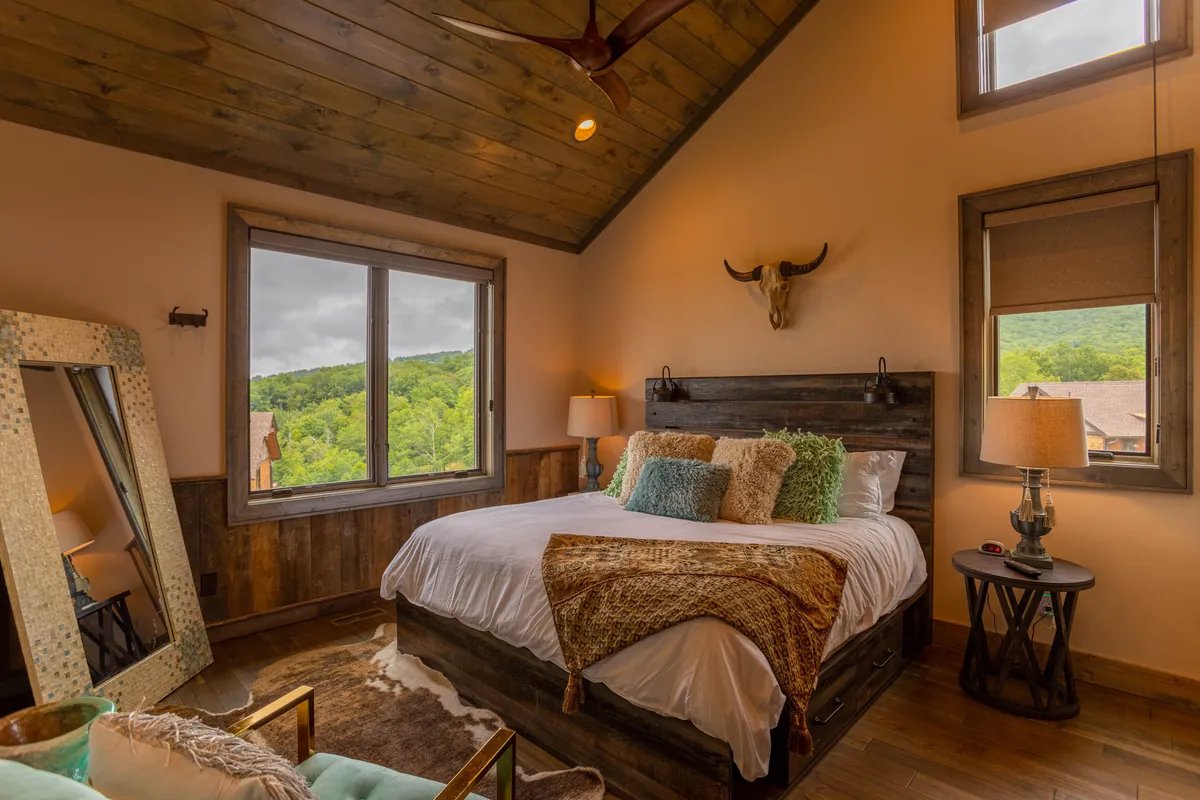 A king suite bedroom with a large bed adorned with decorative pillows, a table lamp on the nightstand, rustic wooden accents, and a spacious window offering a scenic view. A mirror reflects the serene, inviting atmosphere of the room.