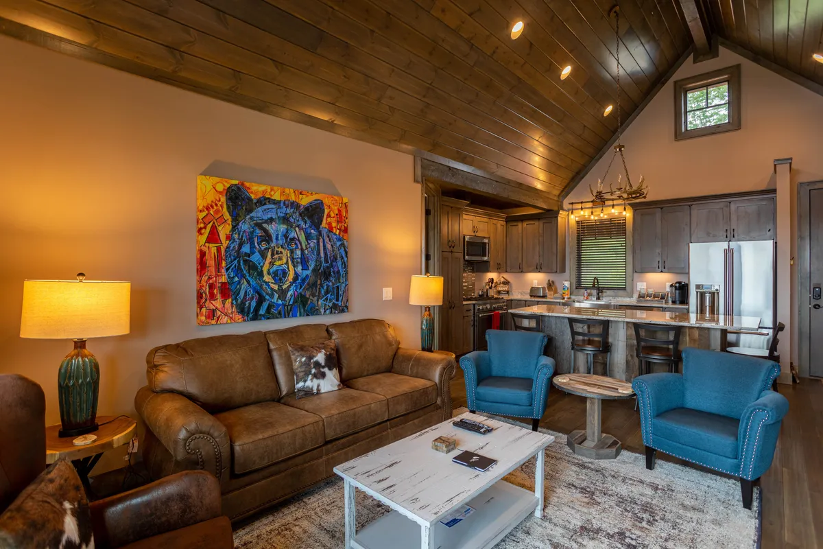 The image presents a vibrant and inviting open-concept area with a comfortable brown sofa, blue accent chairs, a colorful bear painting, and a wooden dining table set for sharing meals. The space feels designed for both relaxation and lively gatherings, with ample seating and artistic touches.
