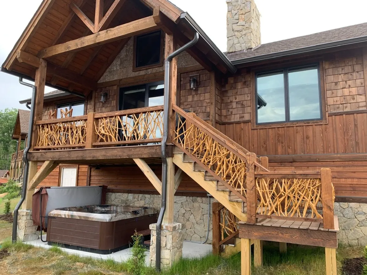This image showcases a wooden balcony and stone foundation, with a hot tub clearly visible alongside an exterior staircase. The vantage point allows viewers to see the relationship between the deck, hot tub, and the surrounding natural landscape, evoking a cozy and inviting atmosphere for social gatherings or relaxation.