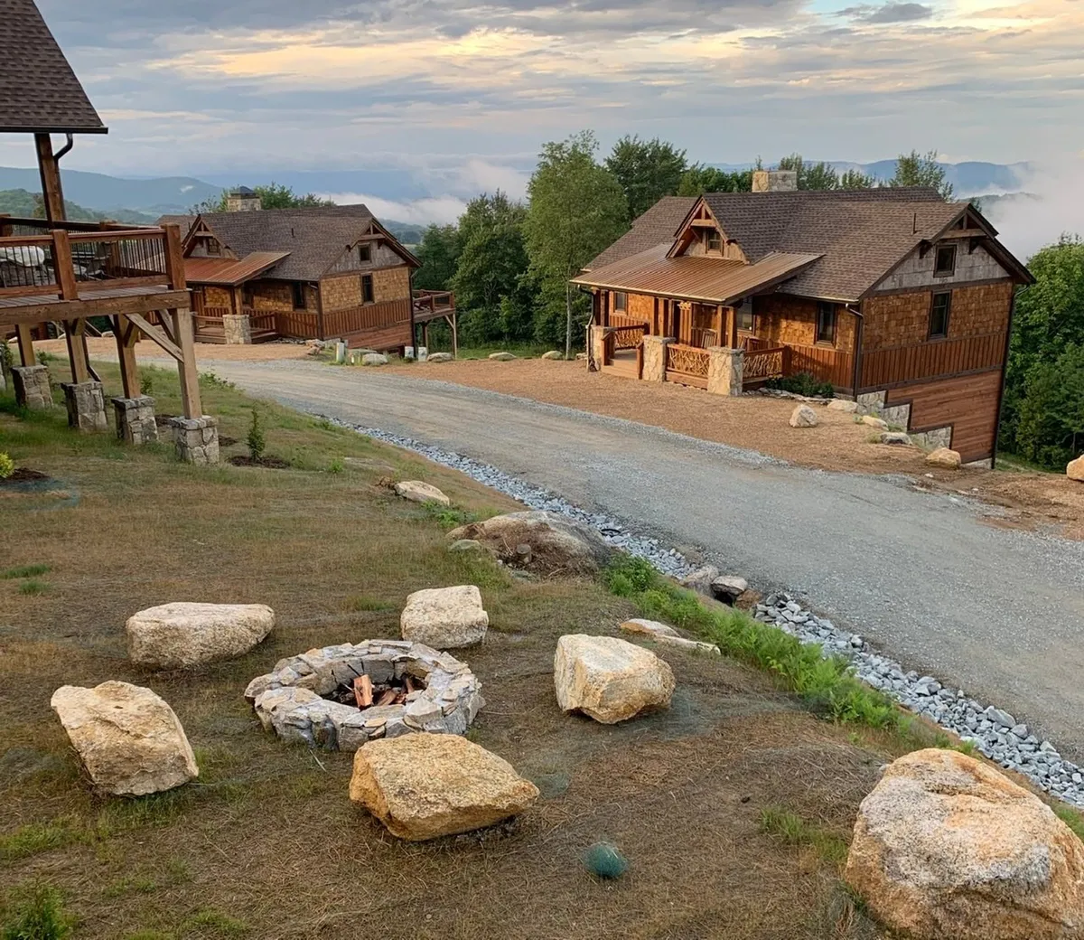 This exterior shot features two rustic cabins flanking a cozy fire pit area on a gravel path. The fire pit is set up for gatherings, with seating arranged for storytelling, s’mores, and stargazing. The surrounding natural landscape adds to the serene vibe.