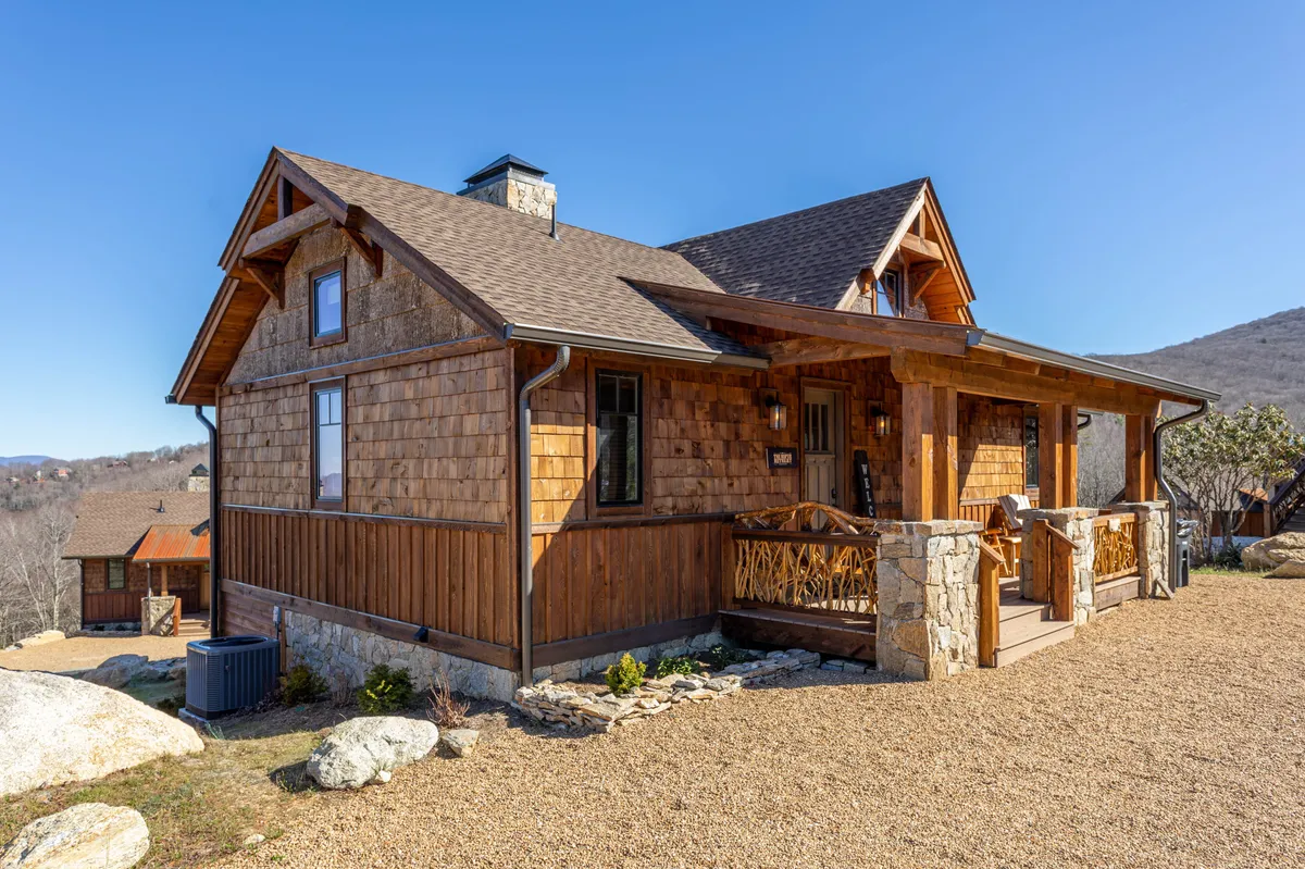 The image showcases the exterior of a charming cabin with a wooden porch, rustic stone accents, and a classic shingled roof, all nestled within a beautifully landscaped garden. The setting invites guests to relax on the porch, enjoy outdoor meals, and take in the expansive views of the surrounding nature.