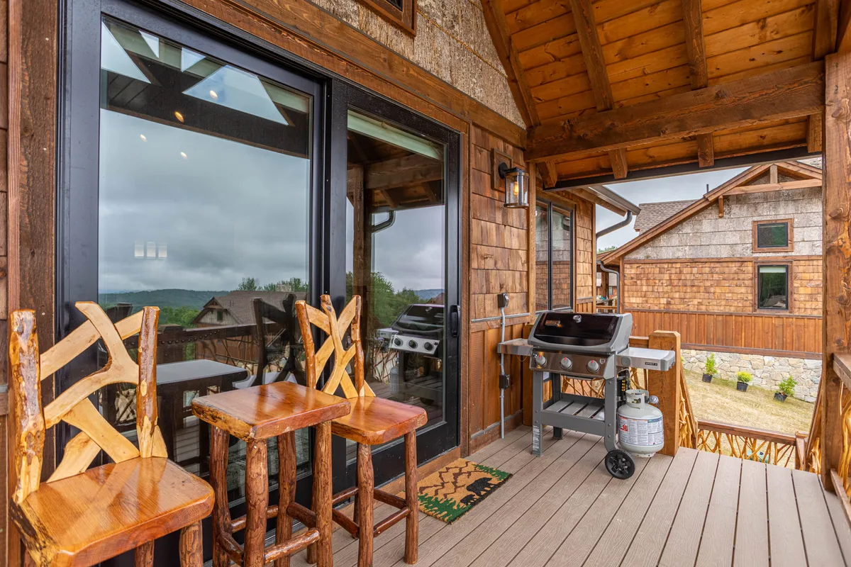This exterior image spotlights a gas grill and wooden bar stools arranged on a deck, set against the backdrop of large windows and a wooden structure. The scene suggests a lively, social atmosphere perfect for barbecuing and enjoying the outdoors with friends or family.